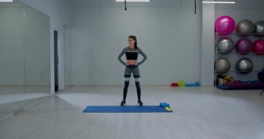 Spor salonundaki mavi bir paspasın üzerinde duran atletik kadın egzersiz rutinine hazırlanırken kafasını sağa sola çeviriyor. Arka planda renkli ağırlık ve egzersiz topları görünür.