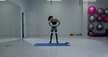 fit bir kadın bir paspasın üzerinde duruyor ve kafa döndürme egzersizi yapıyor. İyi aydınlatılmış bir fitness stüdyosunda idmanına odaklanırken formda olduğunu gösteriyor..