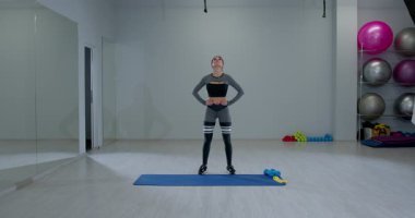 Atletik kadın, parlak bir spor salonunda antrenman rutininin bir parçası olarak başını öne ve arkaya eğiyor. Etrafı sağlık ekipmanlarıyla çevrili ve kararlı..