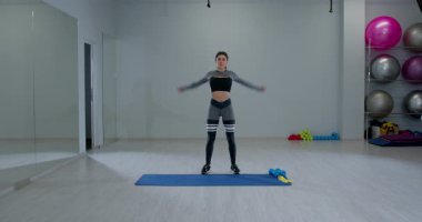 Kararlı atletik kadın, iyi aydınlatılmış bir spor salonunda kol halkaları çiziyor. Etrafı egzersiz ekipmanlarıyla çevriliyken hareketlerine odaklanıyor..