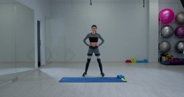 Bayan sporcu, parlak bir spor salonunda antrenmana hazırlanırken mavi bir spor minderinin yanında gülümsüyor. Arka planda renkli fitness ekipmanları görünür.