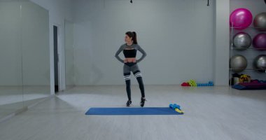 Atletik kadın, parlak bir fitness stüdyosunda tepeden tırnağa sallanıyor. Spor malzemeleriyle çevrili mavi bir paspasın üzerinde dik bir şekilde dengede duruyor..