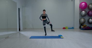 Atletik bir kadın modern bir fitness stüdyosunda dizlerinin yükseldiğini gösteriyor. Alan aktif bir yaşam tarzını destekleyen egzersiz ekipmanları ve aynalar içerir.