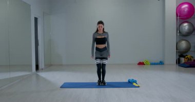 Atletik kadın geniş bir spor salonunda çömelme ve zıplama egzersizi yapıyor. Gücünü ve çevikliğini sergilemeye odaklanmış ve kararlı..