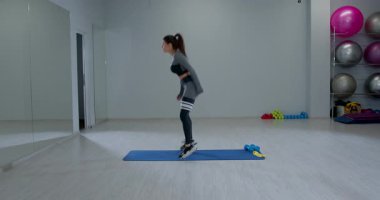 Atletik kadın, gücünü ve çevikliğini göstermek için çömelme hareketi yapıyor. Ayarlar egzersiz ekipmanları ve aynaları içeriyor.