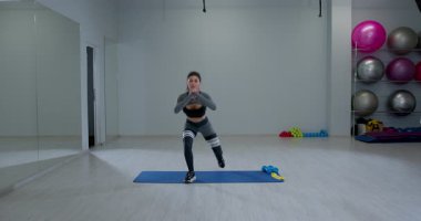 Atletik kadın, parlak bir fitness stüdyosundaki spor minderinin üzerinde ters hamle egzersizi yapıyor. Etrafı spor aletleriyle çevriliyken formuna odaklanıyor..