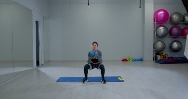 Atletik kadın spor salonunda dambıllarla ters hamleler yapıyor. Çevrede egzersiz paspasları ve ekipmanlar var. Beden eğitimi için parlak ve ferah..