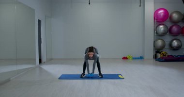 Kendini spora adamış bir kadın geniş bir spor salonundaki mavi bir paspasla kaldırma kombinasyonuna çömeliyor. Alan, arka planda görünen fitness ekipmanlarıyla aydınlık..