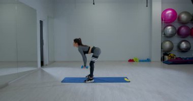 Atletik kadın, parlak bir spor salonunda kaldırma işlemini tamamlamak için çömeldi. Dambıl kullanırken formuna odaklanıyor. Fitness ekipmanları arka planda görünür.