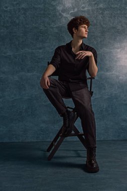 Genç adam siyah gömlekli ve siyah pantolonlu bir sandalyede oturuyor. Düşünceli bir şekilde yukarı bakıyor. Kıvırcık saçları benzersiz tarzına iyi aydınlatılmış stüdyo ortamında katkı sağlıyor..