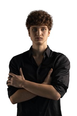 Genç adam kollarını göğsünün önünde kavuşturarak güven içinde ayakta duruyor. Kıvırcık saçları var ve iyi aydınlatılmış stüdyo ortamında siyah bir gömlek giyiyor..