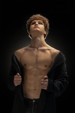 Kıvırcık saçlı genç adam açık siyah bir ceket giyiyor ve göğsünü açıkta bırakıyor. Odaklanmış bir ifadeyle yukarı bakıyor..