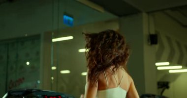 Genç bir kadın, gevşek saçlarıyla koşu bandında koşuyor. Gündüz saatlerinde iyi aydınlatılmış bir spor salonunda çalışmaya odaklanmış ve kararlı. Ortam enerjik ve ilham verici..