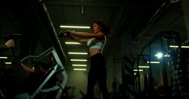 Gevşek saçlı genç bir kadın, bir kettlebell salıncağını çalıştırıyor. Egzersiz rutinine odaklanırken atmosfer enerjiktir. Etrafı çeşitli spor ekipmanlarıyla çevrilidir..