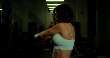 Saçları dökülmüş genç bir kadın jimnastik salonunda omuz gerdirme hareketi yapıyor. Atmosfer, yakınlardaki diğer fitness meraklılarıyla dolu..