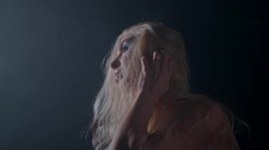 Sarışın bir kadın loş bir alanda durup parmaklarını hafifçe saçlarında gezdiriyor. Yumuşak duman, onun etrafında büyüleyici bir atmosfer yaratır. Olay yerine gizem katar..