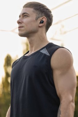 Erkek sporcu, spor sahasında, kulaklıklarla güneş batarken atletik yapısını ve kararlılığını vurgulamaya odaklanmış olarak duruyor..