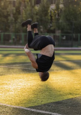 Adam bir futbol sahasında ters takla atıyor. Atletik yeteneklerini sergilerken kulaklık takıyor. Akşamüstü yeşil çimlerle çevrili..
