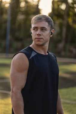 Kulaklıklı bir adam futbol sahasında dikiliyor ve etrafındakilere odaklanıyor. Güneş arka planda batıyor. Egzersiz yaparken sıcak bir atmosfer yaratıyor..