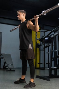 Erkek sporcu, spor salonunda bir halterle çömelme hareketleri yaparken odaklanır. Çevre çeşitli fitness makineleri ve ağırlık ölçüleriyle donatılmıştır..