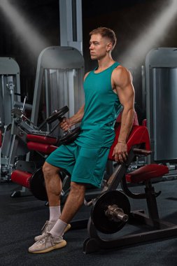 İyi aydınlatılmış bir spor salonunda ağırlık kaldırırken odaklanmış bir atlet kısa bir ara verir. Kurulumda modern ekipmanlar yer alıyor ve güç eğitimi ve uygunluğu vurgulanıyor.