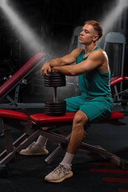 Atletik bir insan bir spor salonunda, elinde bir dambılla oturup dinlenir. Ortam, antrenmanlara adanmış çeşitli spor aletleriyle dolu bir fitness salonu..