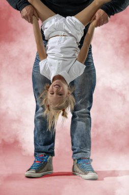 Babası onu kollarından tutarken küçük sarışın bir kızın neşeyle baş aşağı asılı durduğu bir stüdyoda. Çevreleri parlak bir arka planla çevrili. Eğlenceli bir oyun ortamı yaratıyorlar..