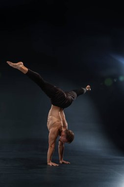 Kaslı adam bir elinde denge kurarken amuda kalkıp güç ve denge gösterisi yapıyor. Karanlık arka plan, parlak spot ışıklarıyla çelişiyor..