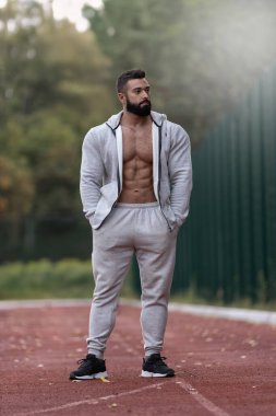 Atletik kıyafetler giymiş kaslı bir adam koşu pistinde duruyor. Zindelik yolculuğunu vurgulayarak odaklanmış ve rahatlamış görünüyor. Arkaplanda ağaçlar ve açık bir gökyüzü var..