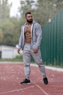 Atletik adam spor sahasında poz veriyor. Gücünü ve uygunluğunu gösteriyor. Gri bir eşofman giyiyor ve ağaçların yeşil zeminine karşı güçlü bir kas yapısı gösteriyor..