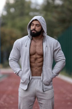 Atletik adam spor sahasında açık gri kapüşonlu ve uyumlu pantolonlu duruyor. Forma ve sağlığa olan bağlılığını gösteren güçlü bir fiziği var..