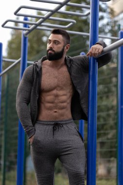 Spor sahasında spor aletlerinin yanında duran kaslı adam spor kıyafetleri giyerken karın kaslarını gösteriyor. Ortam ağaçlarla ve parlak gökyüzüyle dolu..