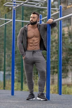 Atletik bir adam, öğleden sonra güneşinin tadını çıkarırken güçlü fiziğini ve kararlılığını sergileyen barların yanındaki spor sahasında güven içinde poz verir..