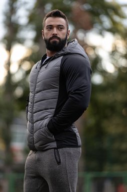 Gri yelek ve eşofman giyen atletik bir adam elleri spor sahasında cebinde duruyor. Batan güneş, akşam ışığında etrafında sıcak bir atmosfer yaratıyor..