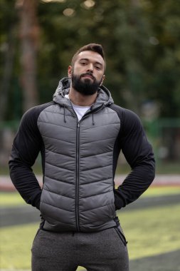 Sporcu bir adam spor sahasında montlu bir ceket giyer ve kameraya kendinden emin bir şekilde bakar. Ayarda canlı bir yeşil alan ve arka planda ağaçlar yer alıyor..