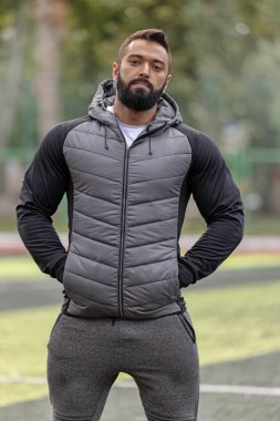 Sporcu adam spor sahasında şık bir ceket ve pantolon giyer. Odaklanmış ve kendinden emin görünüyor. Muhtemelen güneşteki alıştırmalar arasında mola veriyor..