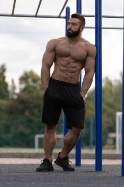 Güçlü bir atletik adam spor sahasında kendinden emin bir şekilde duruyor. Etkileyici kaslı bir fiziği var ve spor yapmak için giyinmiş. Forma ve eğitime olan bağlılığını vurguluyor..