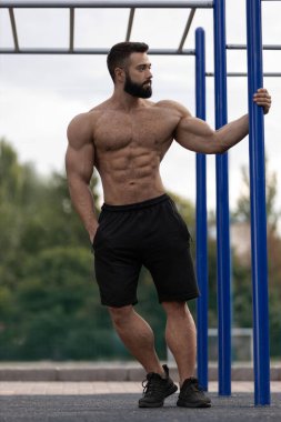 Atletik bir adam spor sahasında, barın karşısında dinlenirken kaslı vücudunu sergiliyor. Çevre, dışarıda çalışmak ve spor yapmak için idealdir..
