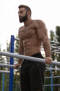 Kaslı bir adam, akşam güneşinin sıcacık parıltısında gücünü ve atletizmini gösteren bir spor sahasında paralel barlarda dalış yapar. Zindeliğe olan bağlılığı çok açık..