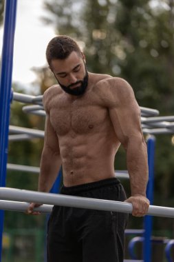 Kas yapısı olan atletik bir adam spor sahasında spor yapıyor. Egzersiz ekipmanları ve yeşilliklerle çevriliyken yatay bir çubukta gücünü gösteriyor..