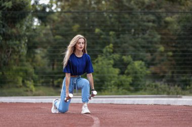 Kız sokakta diz çökerken dambıllarla çalışıyor. O, güç eğitimine odaklanır. Sahne, arka planda ağaçlar ve açık bir gündüz gökyüzünü gösteriyor..