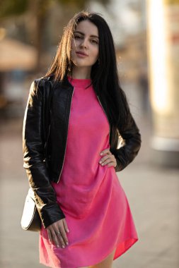 Kız, açık pembe bir elbise ve siyah bir ceket giyerek bir şehir caddesinde güven içinde poz veriyor. Güneş ışığı arka planı aydınlatır ve canlı bir atmosfer yaratır..