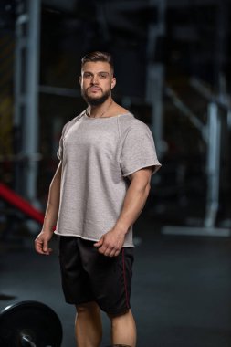 Adam odaklanmış ve pazı kıvırmak için halteri kaldırmaya hazır. Ekipmanlarla dolu bir spor salonunda. Akşam oldu ve o idman kıyafetlerini giymiş..