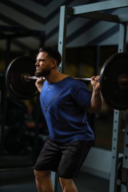 Adam ağır bir halterle çömelme egzersizi yapıyor. Jimnastik salonunda. Etkinlik onun güç eğitim rutininin bir parçası. Gündüz vakti..