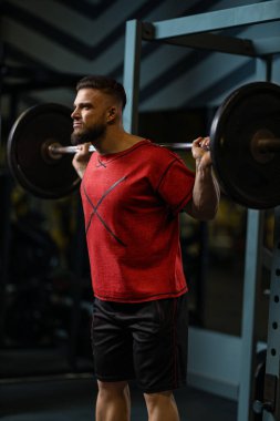 Adam spor salonunda omuzlarında halterle çömelme hareketleri yapıyor. Ağırlık kaldırırken formuna odaklanıyor. Spor salonu çeşitli spor aletleriyle donatılmıştır..