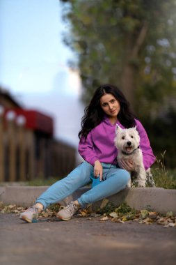 Kız West Highland White Terrier 'ını tutarken bir patikada dinleniyor. Çevreleri yeşil ağaçlar ve açık bir gökyüzüyle çevrilidir. Gündelik ve neşeli bir an..