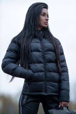 Esmer kadın siyah ceketli ve siyah bir sırt çantası taşıyan dışarıda duruyor. Gökyüzü bulutlu görünüyor. Hareketsiz dururken düşünceli bir şekilde yana bakıyor..