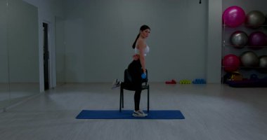 Yoga minderinin üzerinde, geniş spor salonunda küçük bir ağırlık ve sandalyeyle duran formda bir kadın antrenör.