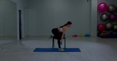 Formda bayan sporcu, geniş spor salonunda aynalarla yoga minderinin üzerinde mavi ağırlığı kaldırıyor.