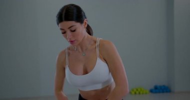 Kız spor salonunda egzersiz yaparken öne eğiliyor. Etrafında çeşitli ağırlıklar olan açık bir alanda rutinine odaklanıyor. Işık basit bir atmosfer yaratır..